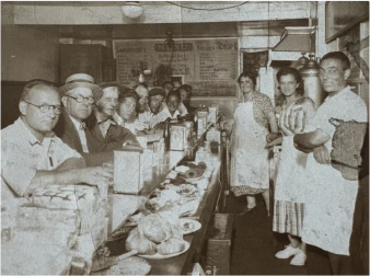A faded, sepia-toned photograph captures a bustling diner scene. On the left, a long counter is lined with several male customers, most wearing glasses and looking towards the right. One man in the foreground on the left wears a light-colored shirt and glasses, leaning on the counter. Various food items are displayed on the counter in front of the customers. On the right side of the image, two women and one man, all appearing to be staff members, stand behind the counter. The woman closest to the foreground wears a patterned dress and a white apron, smiling towards the camera. Behind them, a menu board with "SANDWICHES," "SALADS," and "CHOPS" written at the top is visible. The lighting is dim, suggesting an indoor setting.