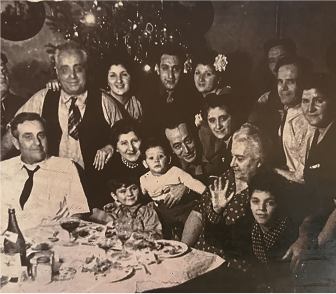 A sepia-toned, black and white photograph shows a large family gathered around a table, likely celebrating. The table in the foreground holds a bottle, glasses, and plates with food. A diverse group of adults and children are present, some smiling and looking towards the camera. One older woman in the center-right is waving towards the viewer. A man to her left holds a young child. Another young child sits at the table in the lower left. In the background, what appears to be festive decorations or a Christmas tree with ornaments is visible. The overall atmosphere suggests a warm, convivial family gathering.