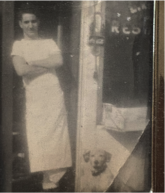 A faded, sepia-toned photograph shows a young man standing with his arms crossed, wearing a light-colored t-shirt and a long apron. He is standing in a doorway or entrance. Below and slightly to his right, a small, light-colored dog with dark eyes is looking directly at the camera. The background behind the dog is dark, possibly the interior of a building or a shaded area.