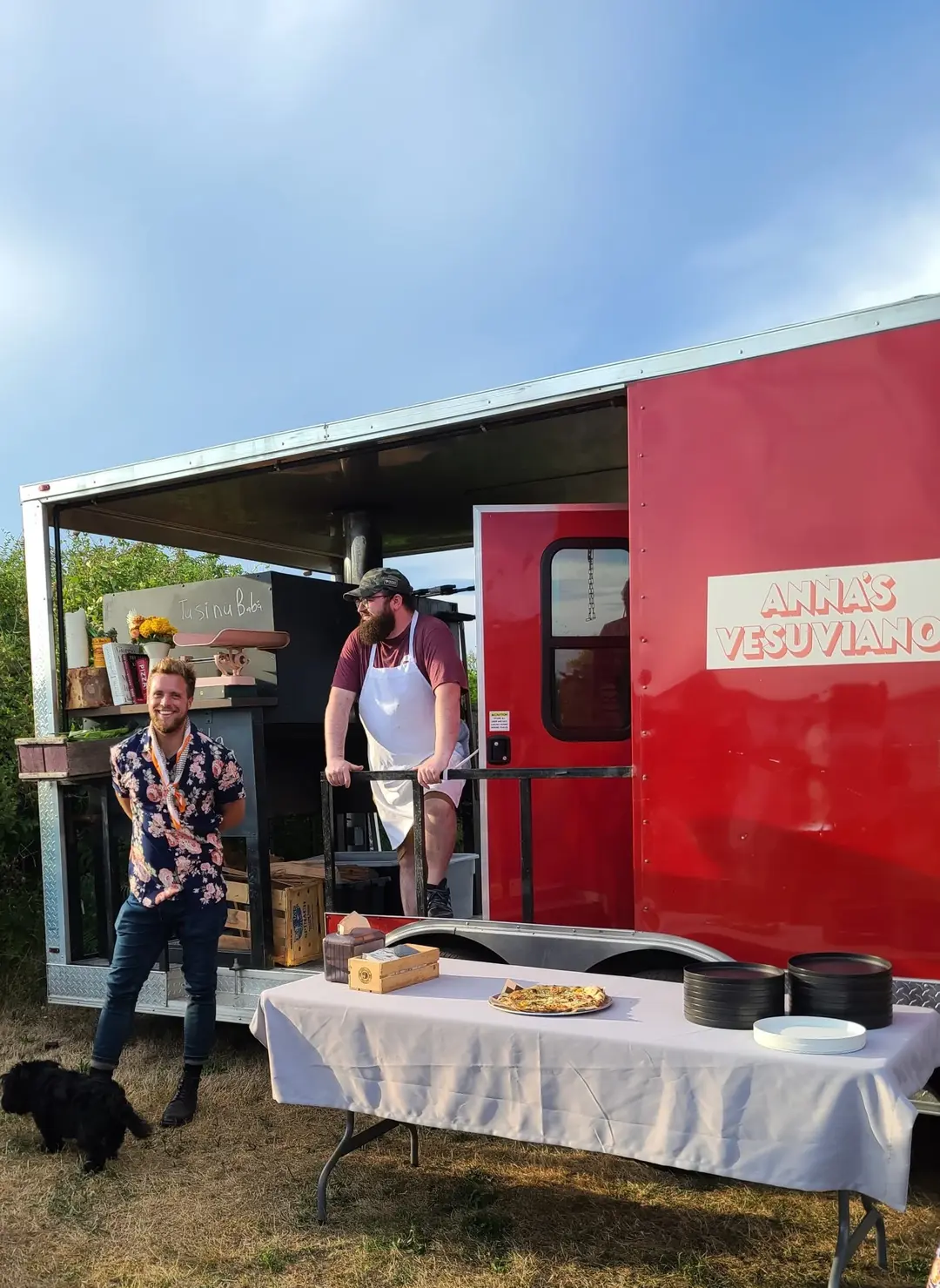 A bright red food truck, possibly a pizza truck, is open for business outdoors. The side of the truck has "ANNA'S VESUVIANO" written in a light, possibly orange or yellow, font. A man with a beard and a white apron stands inside the truck, looking out. To the left, a younger man with short hair and a floral shirt stands next to a small black dog, smiling. In front of the truck, there's a table with a white tablecloth, holding plates, bowls, and a cooked pizza. The setting appears to be a sunny day with clear skies and green grass.